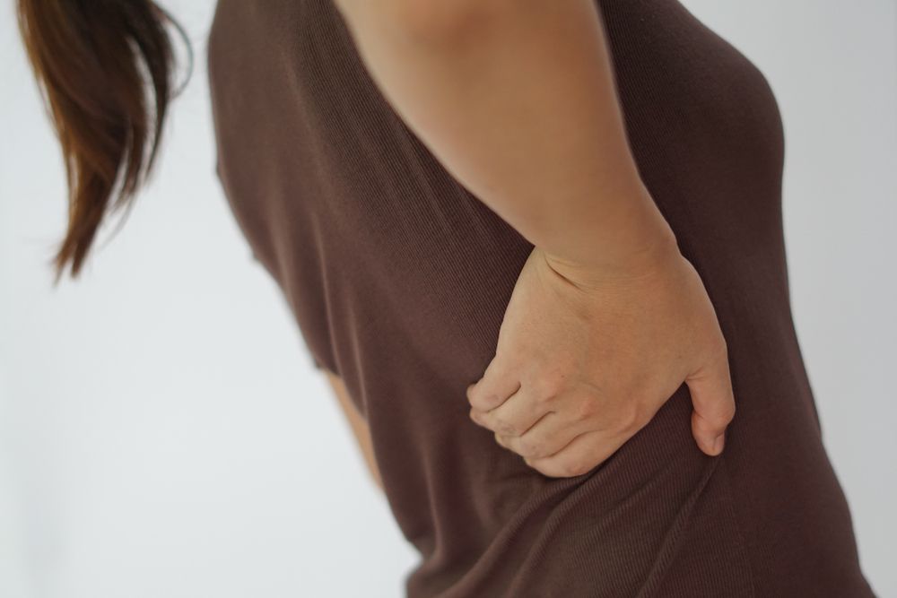 Mujer sujetándose la espalda baja, top marrón, fondo blanco.