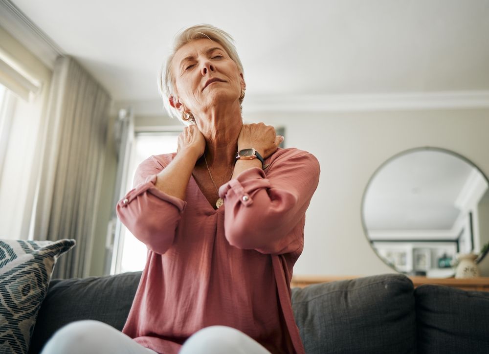 Mujer en un sofá con las manos en el cuello, indicando dolor cervical. Interior, con camisa rosa.