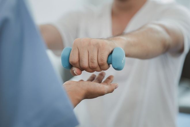 Persona sosteniendo una mancuerna azul, ejercitando el brazo con la ayuda de alguien con uniforme azul.