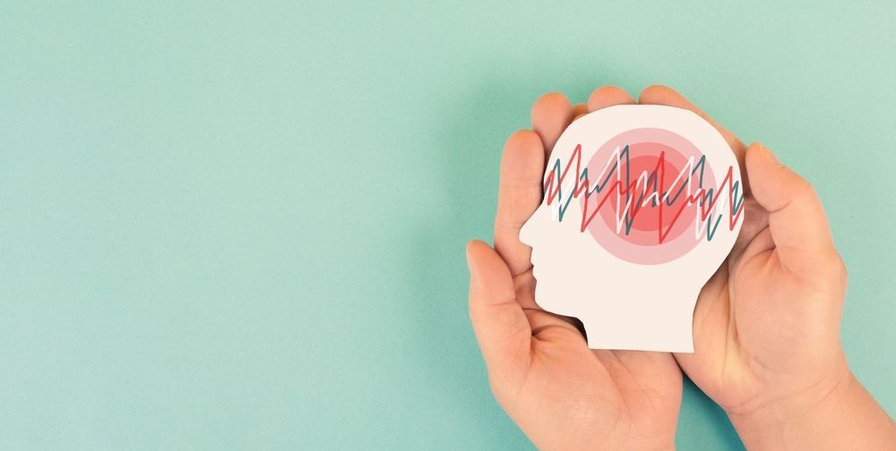 Manos sosteniendo una cabeza recortada de papel con líneas rojas onduladas y un círculo rojo en el interior, sobre un fondo turquesa.