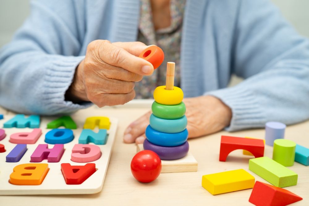 Persona mayor apilando anillos de colores en una clavija de madera, jugando con juguetes en una mesa.
