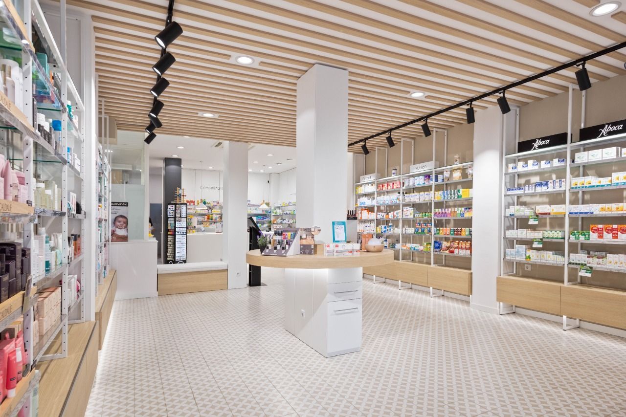 Interior de una farmacia luminosa con filas de estantes llenos de productos, suelo de baldosas blancas y techo de madera.