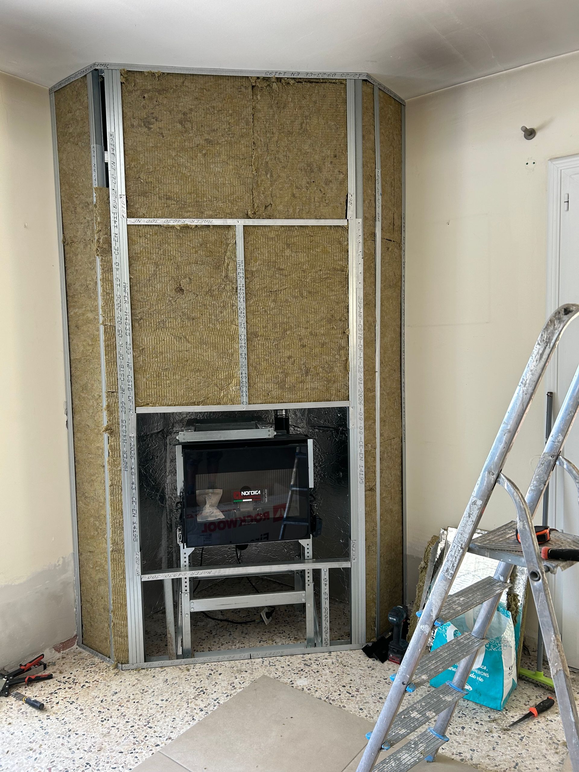 Mur de cheminée partiellement fini avec isolation apparente et insert de cheminée, échelle dans une pièce en rénovation