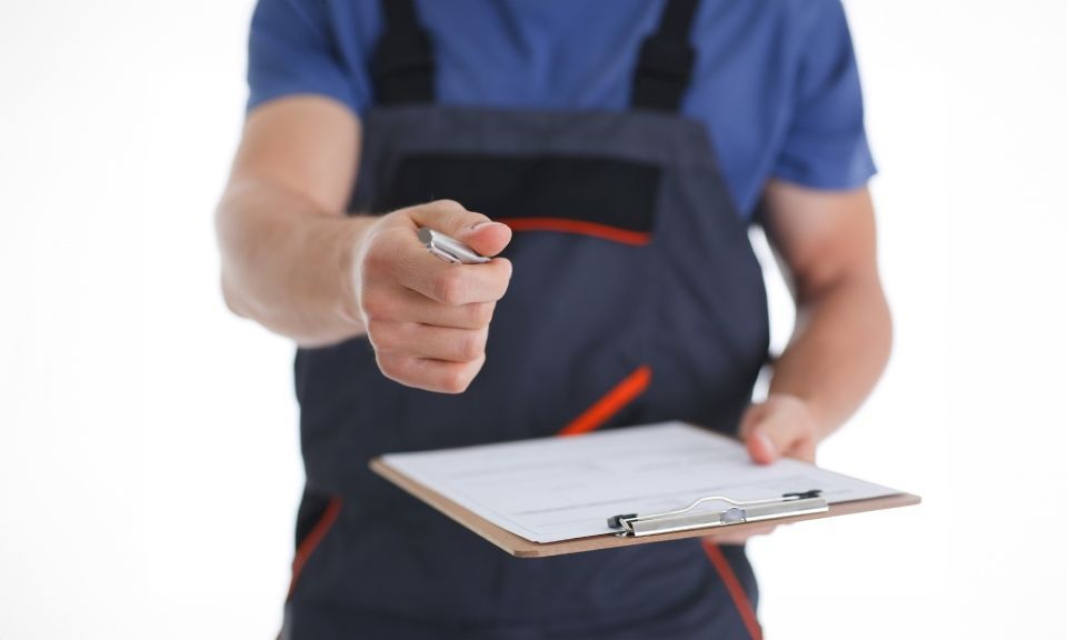 Une personne en salopette de travail bleue tend un stylo et un bloc-notes pour une signature sur fond blanc.