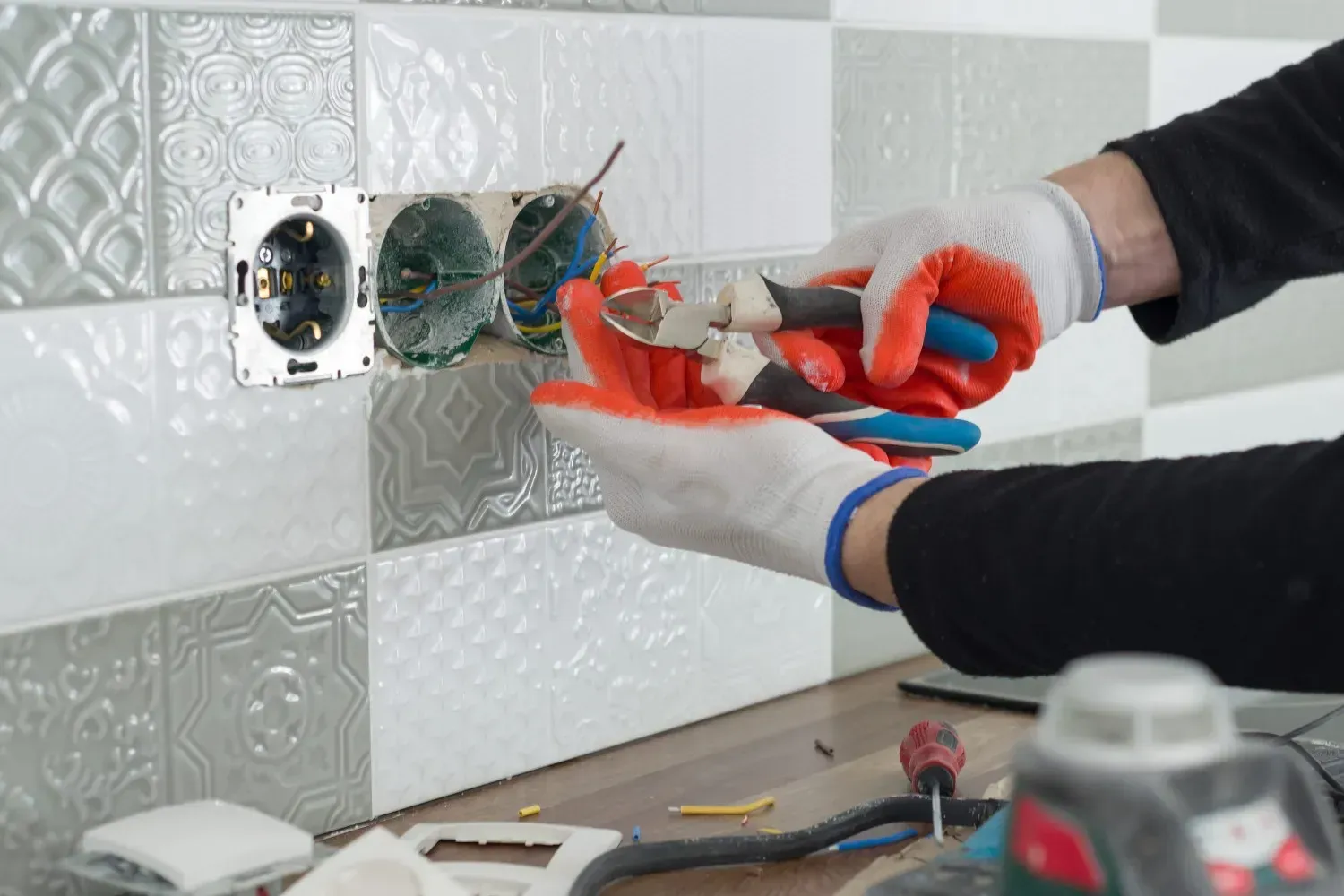 Un electricista con guantes naranjas utiliza unos alicates para cortar los cables eléctricos de un enchufe empotrado en una pared de azulejos.