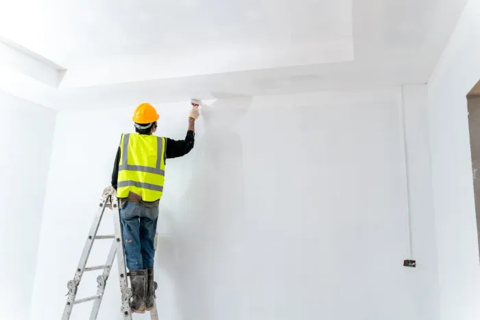 Un trabajador con camisa blanca y guantes azules utiliza una herramienta para aplicar sellador al borde del voladizo blanco de un edificio.