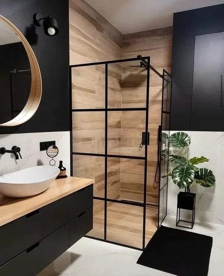 Un baño moderno con tocador negro, lavabo sobre encimera, espejo redondo y ducha a ras de suelo con paneles de vidrio negro con rejilla.