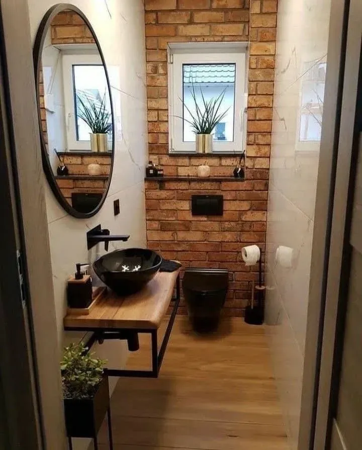 Un moderno aseo de estilo industrial con una pared decorativa de ladrillo visto, tocador de madera, lavabo negro sobre encimera y espejo redondo.