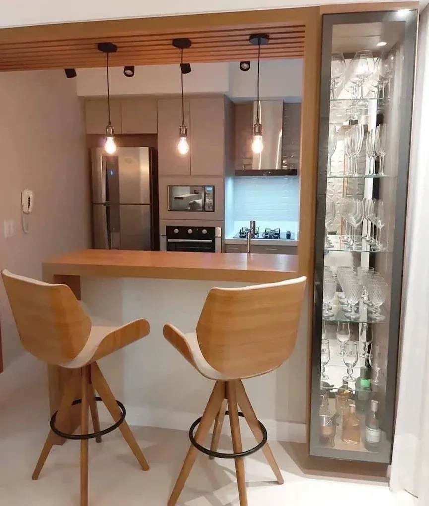 Una zona de barra de cocina con dos taburetes de madera clara, tres lámparas colgantes, un panel de techo de madera y una vitrina alta de cristal.