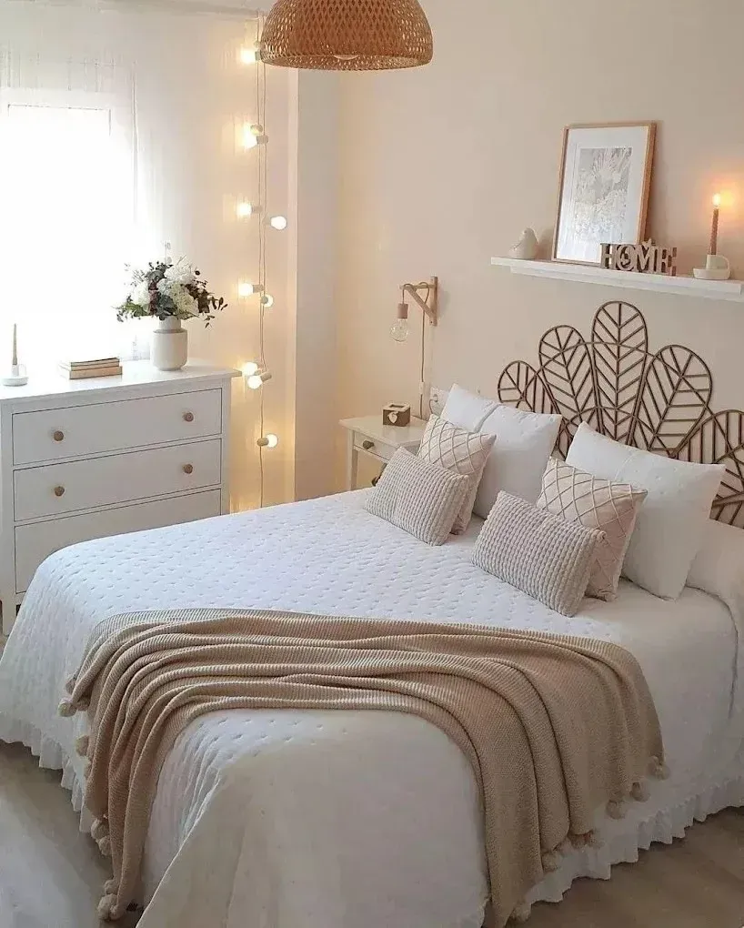 Un acogedor dormitorio en tonos crema con cabecero de ratán con forma de pavo real, ropa de cama blanca con una manta beige y luces de hadas cálidas.