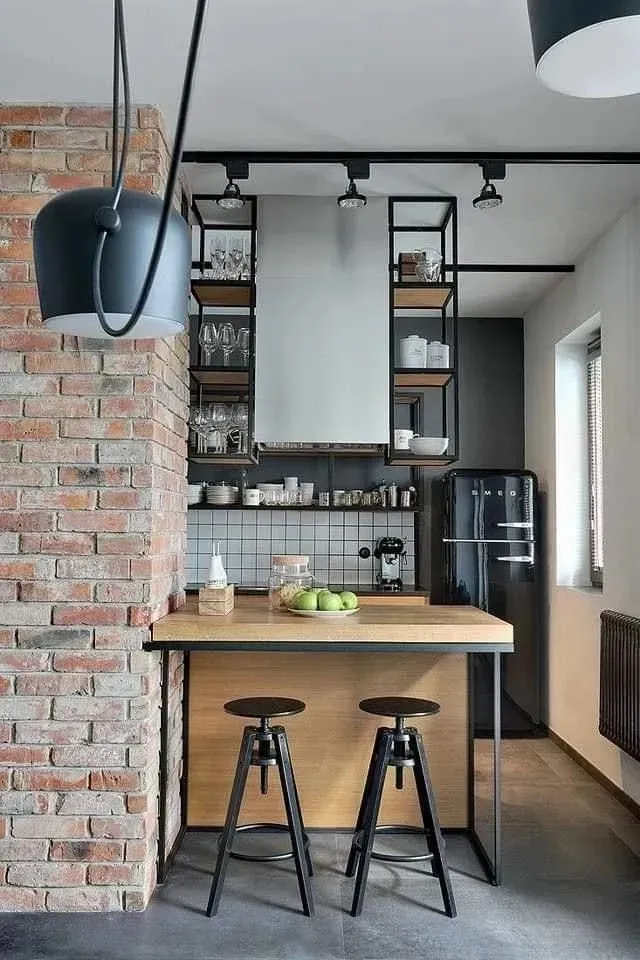 Una pequeña cocina de estilo industrial con una barra de desayuno de madera, taburetes de metal negro y estantes abiertos contra una pared de ladrillo.