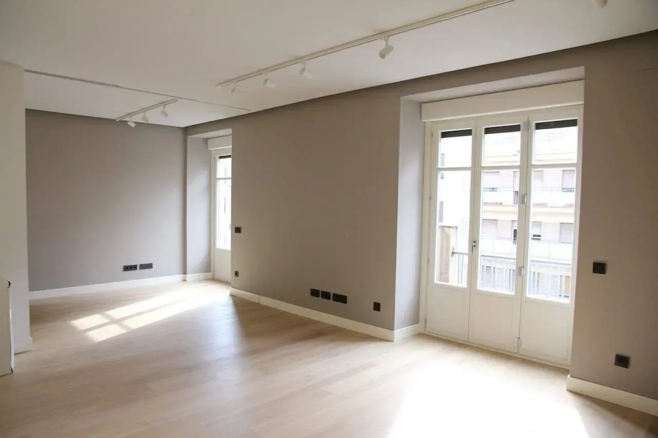 Una habitación vacía y bien iluminada, con paredes de color beige, suelos de madera clara, iluminación empotrada y un conjunto de puertas dobles de cristal.