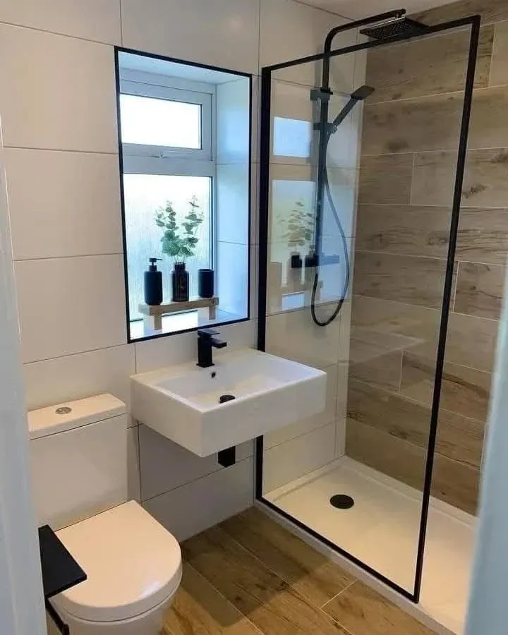 Un baño moderno con lavabo blanco montado en la pared, inodoro y ducha a ras de suelo con marco negro y azulejos que imitan la madera.