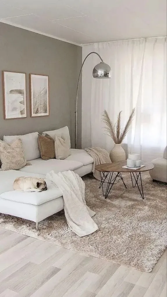 Una acogedora sala de estar con un sofá blanco, cojines de tonos neutros, una alfombra de pelo largo, una lámpara de pie cromada y un jarrón con ramas secas.