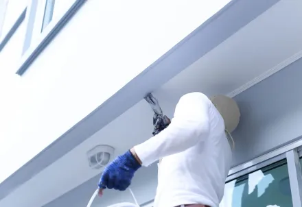 Un trabajador con camisa blanca y guantes azules utiliza una herramienta para aplicar sellador al borde del voladizo blanco de un edificio.