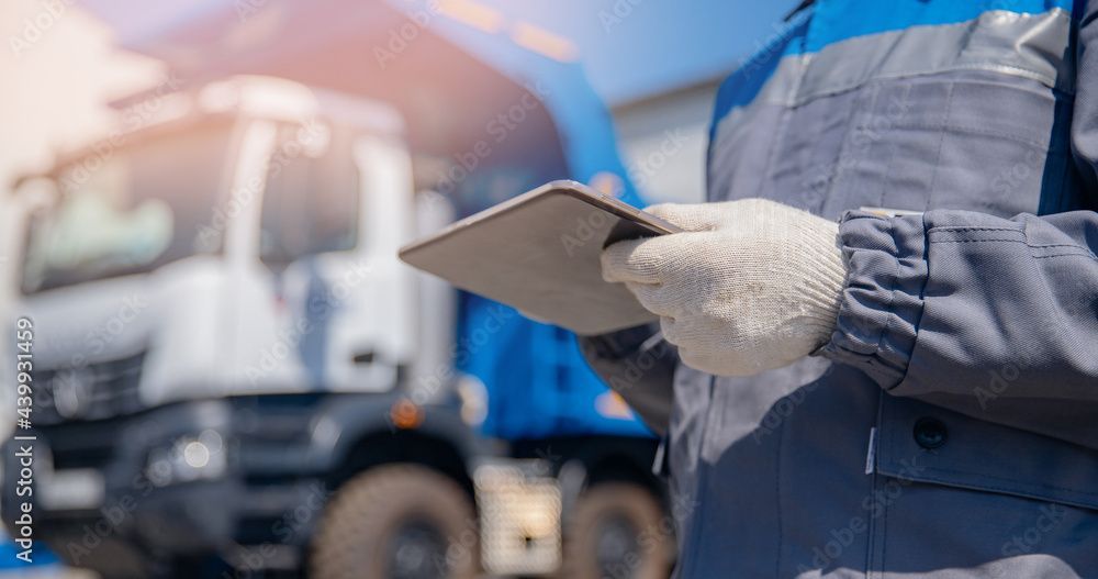 Ein Arbeiter in blauer Arbeitskleidung und Handschuhen hält ein Tablet vor einem LKW im Freien.