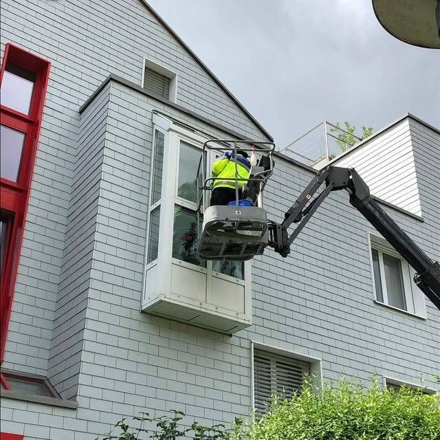 Ein Arbeiter repariert in einem Aufzug ein Fenster an einem grauen, mit Schindeln gedeckten Gebäude.