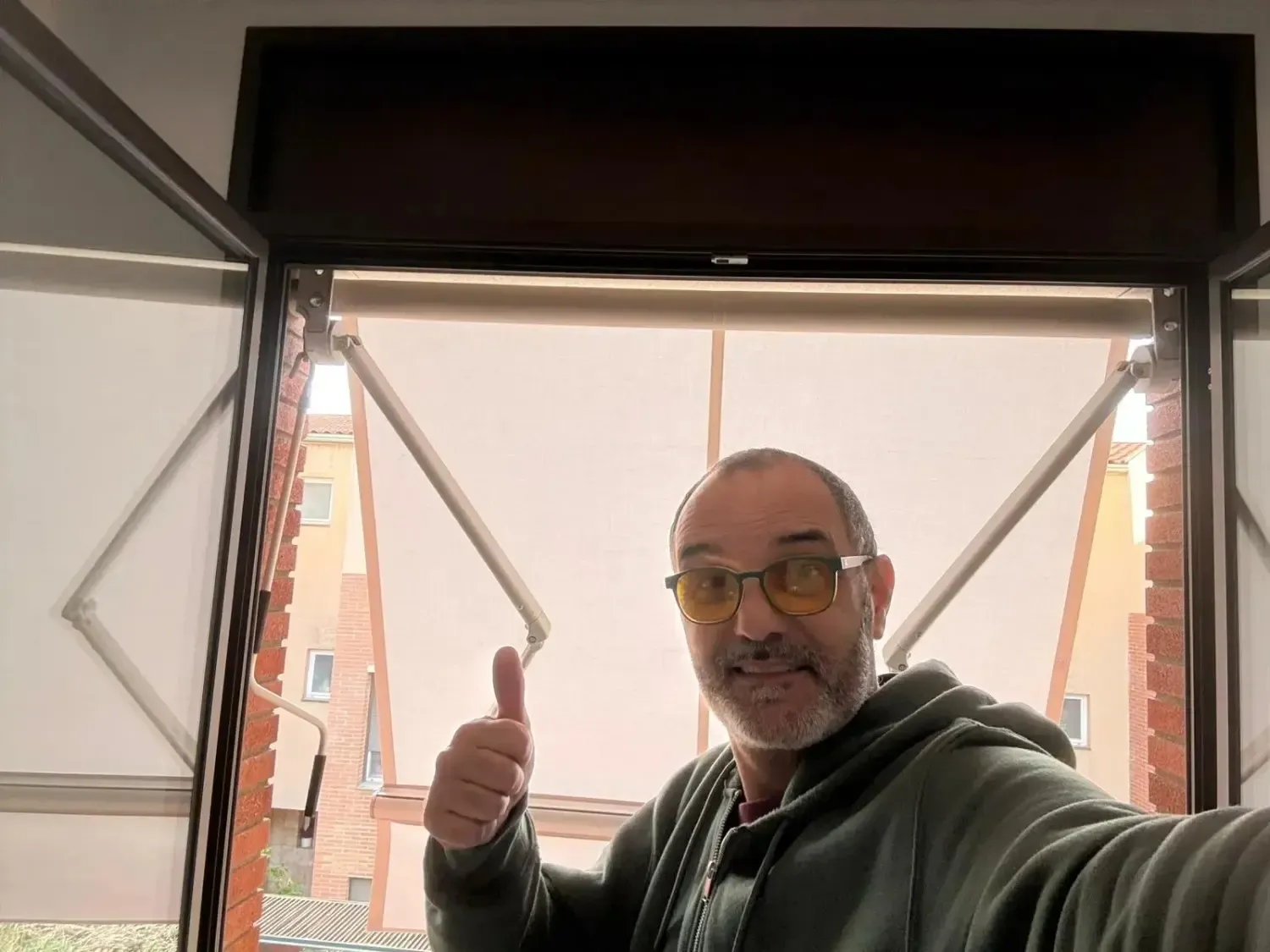 Un hombre con gafas amarillas hace un gesto de aprobación con el pulgar frente a una ventana con las persianas cerradas, vista al exterior.