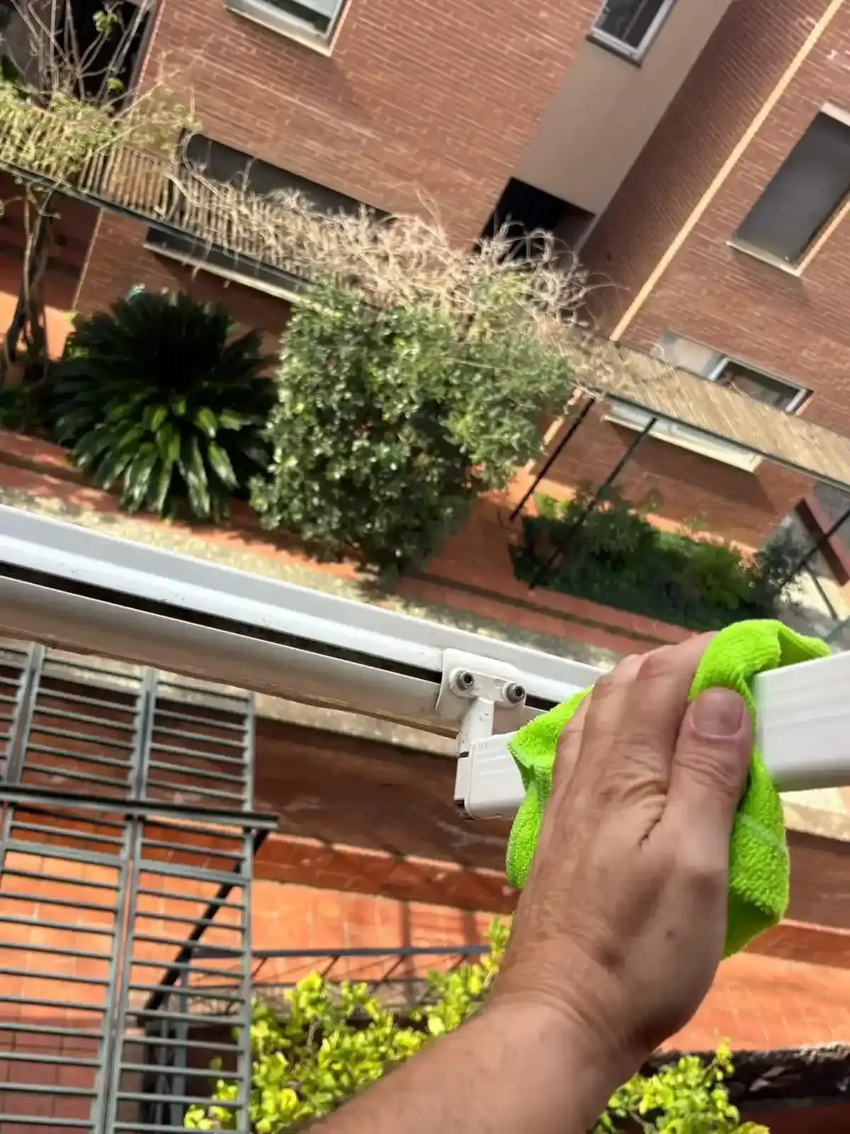 Mano con un paño verde limpiando una repisa blanca, con un edificio y plantas al fondo.