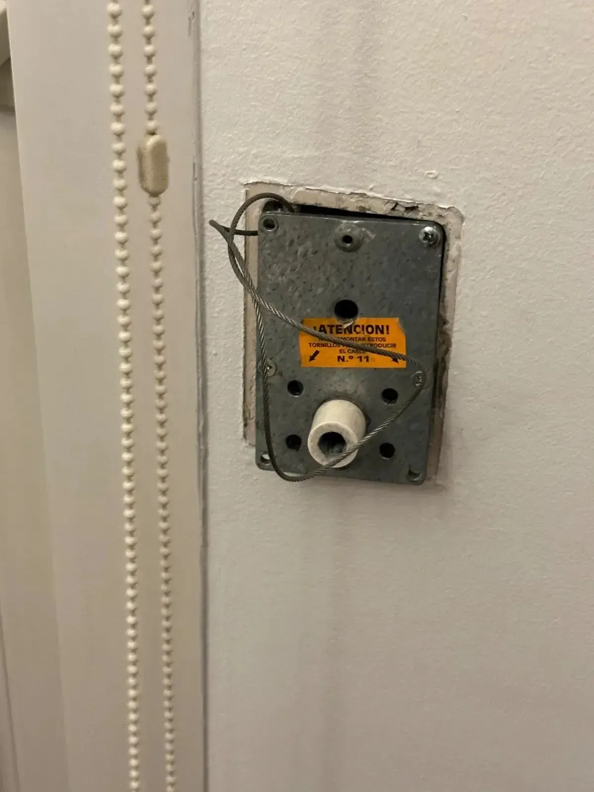 Caja eléctrica de metal sobre una pared blanca con cableado y un cilindro blanco adjunto; al lado de un cordón de cuentas verticales.