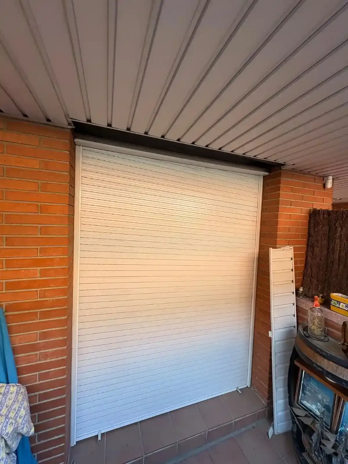 Persiana enrollable blanca que cubre una abertura de ladrillo debajo de un techo corrugado.