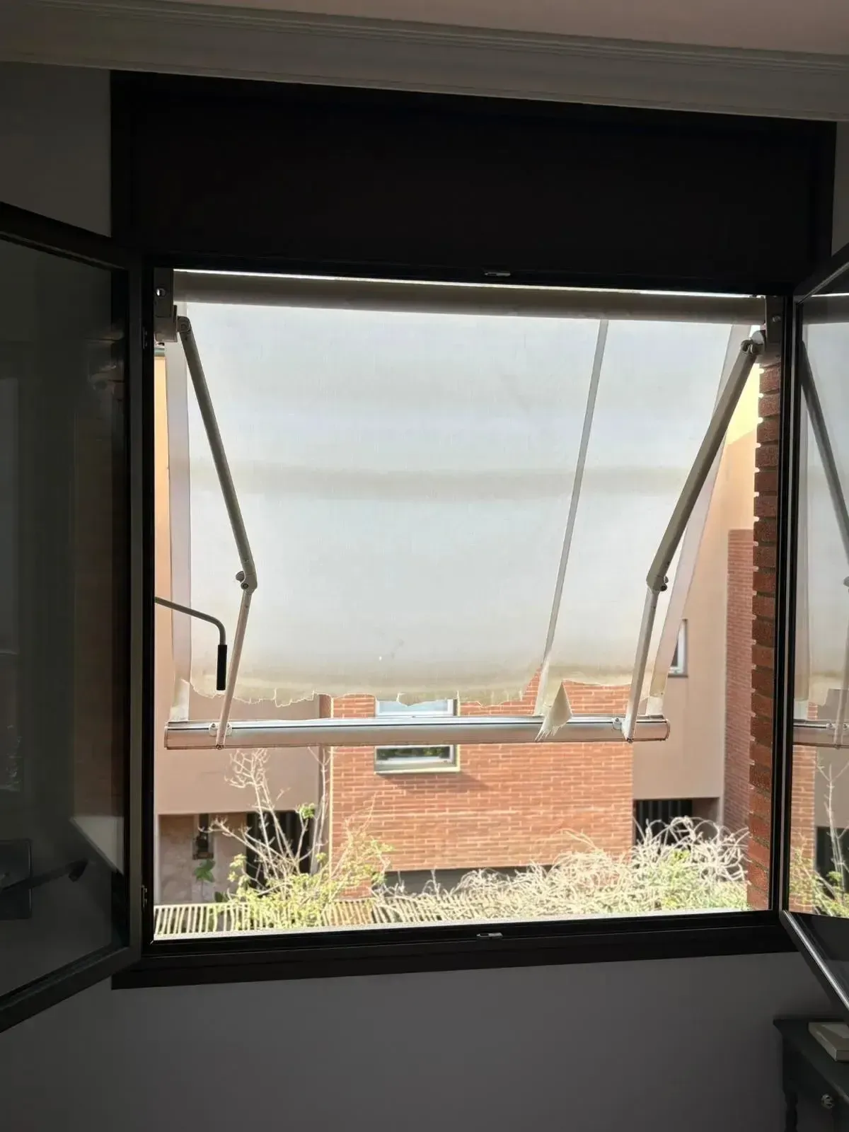 Ventana abierta con persiana blanca parcialmente bajada, revelando un edificio de ladrillo.