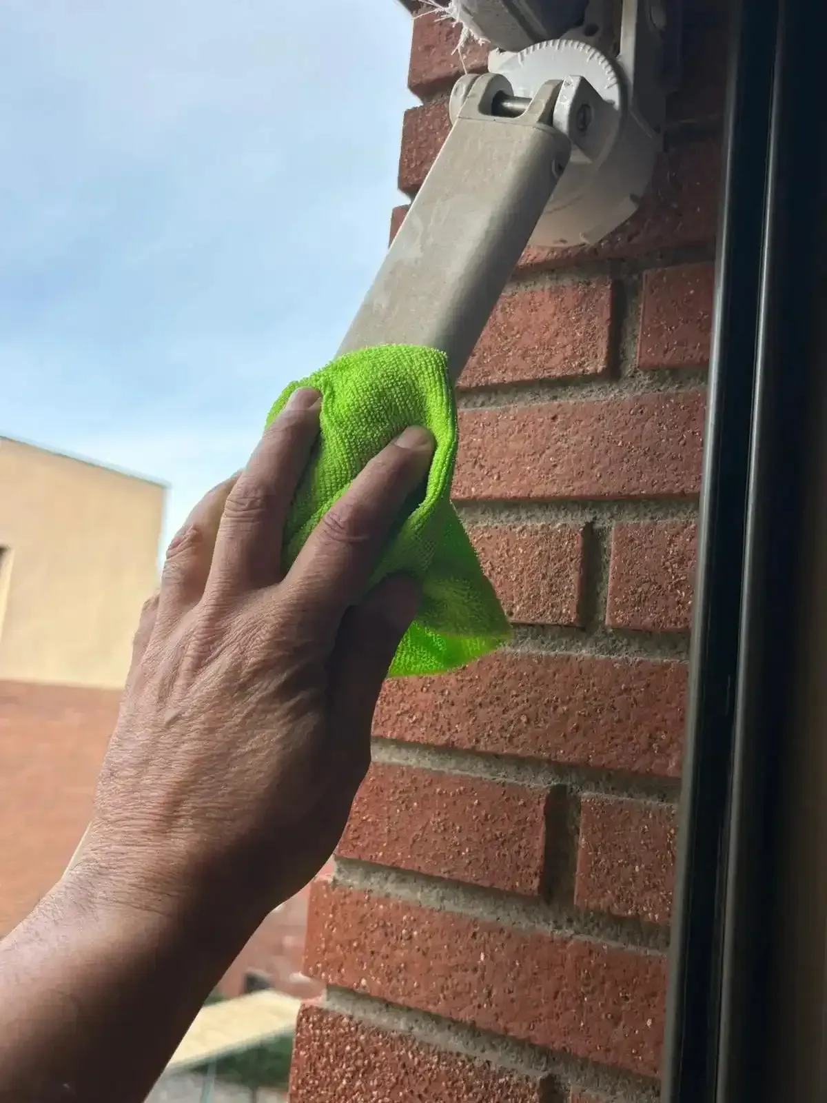 Mano limpiando un soporte blanco en una pared de ladrillo rojo con un paño verde.