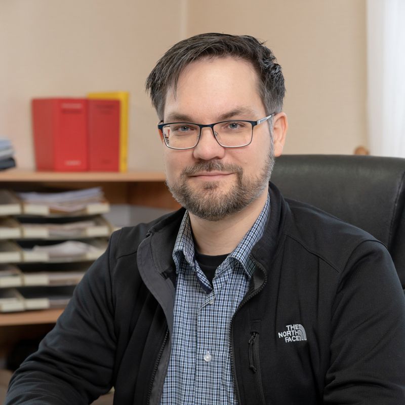 Ein Mann mit Brille und schwarzer Jacke sitzt an einem Schreibtisch.
