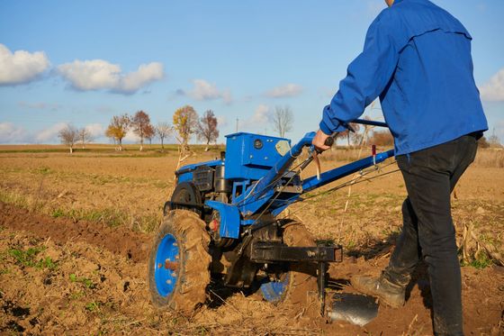 Une personne conduit un tracteur bleu à conducteur marchant dans un champ labouré sous un ciel dégagé.