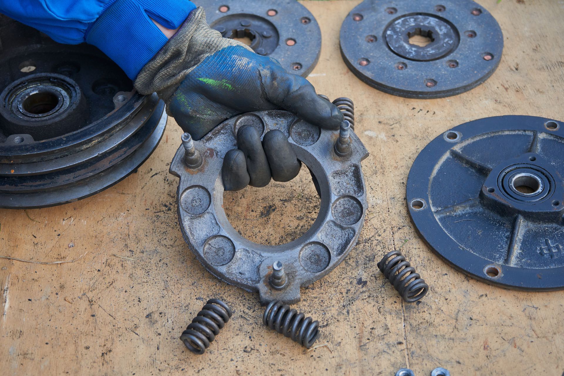 Des mains gantées tiennent un moyeu de roue métallique usé, entouré de pièces circulaires bleues et de ressorts, sur un établi.