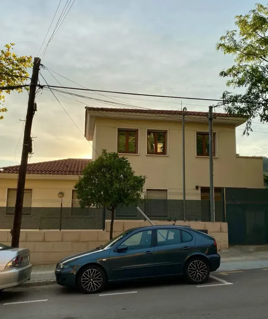 Un coche azul oscuro aparcado en la calle frente a una casa beige de dos plantas con tejado de tejas al anochecer.
