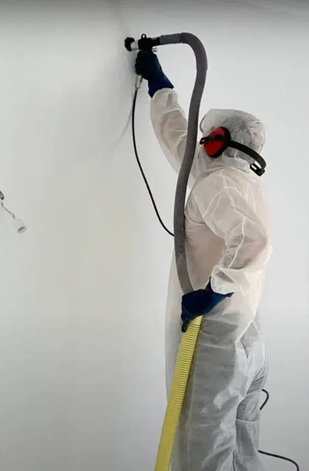 Una persona con ropa protectora blanca y gafas de seguridad utiliza una lijadora de pared conectada a una manguera