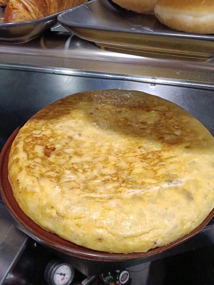 Una tortilla española redonda sobre un plato oscuro, con productos horneados en el fondo.