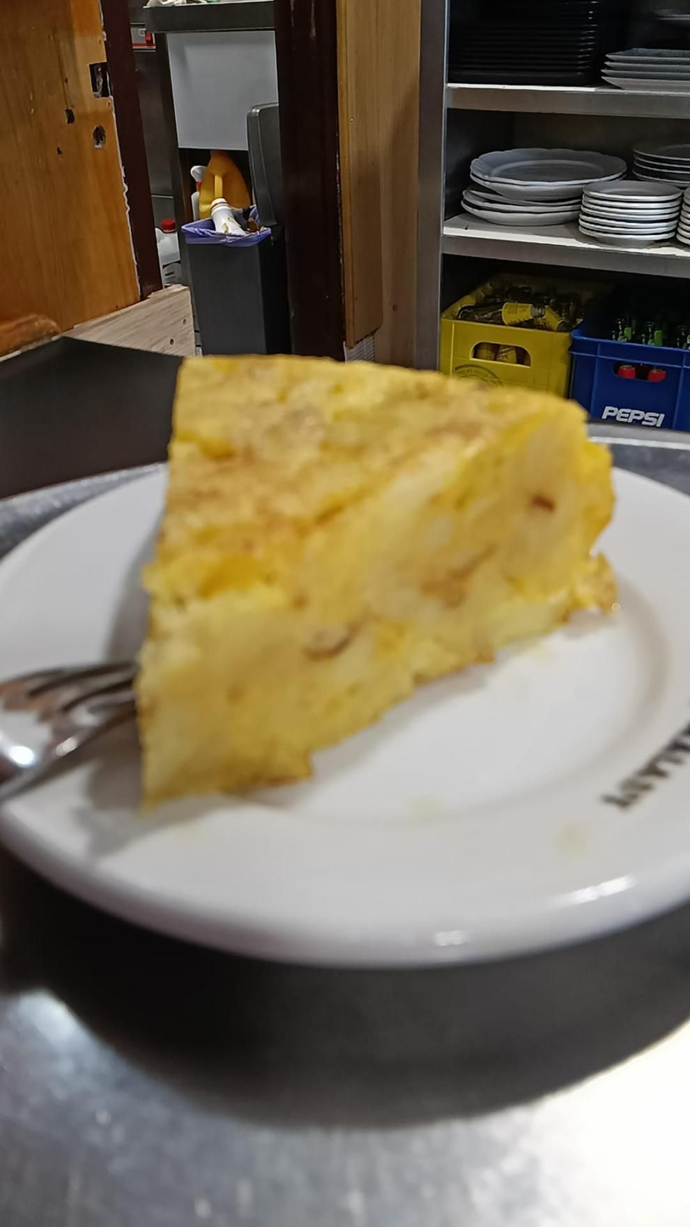 Rebanada de tortilla de patatas en un plato blanco con un tenedor, entorno de restaurante.