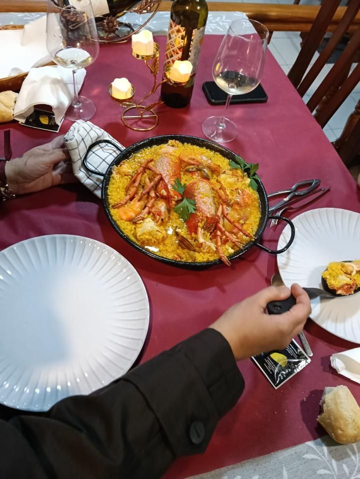 Paella en una sartén sobre un mantel rojo con platos y copas de vino, manos buscando comida.