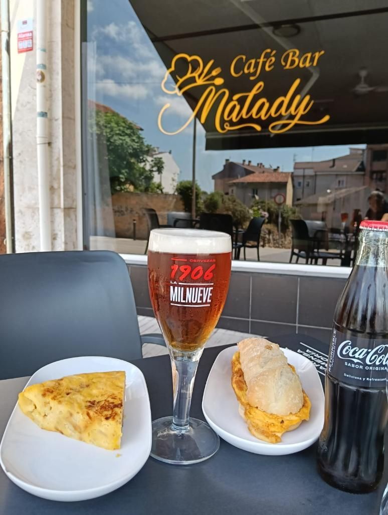 Mesa de café al aire libre con cerveza, tortilla, sándwich y botella de Coca-Cola frente a una ventana.