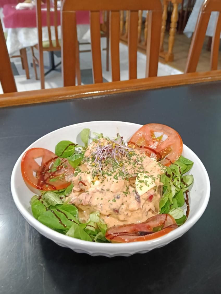 Ensalada en un tazón blanco: tomates, lechuga y cobertura cremosa con hierbas. Mesa y sillas de restaurante al fondo.