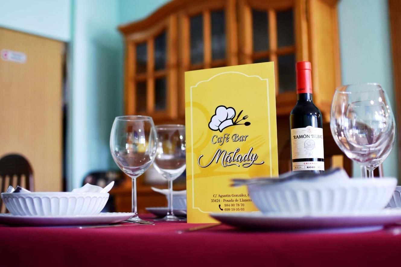 Puesta de mesa en un restaurante: botella de vino, menú, copas y platos sobre un mantel rojo.
