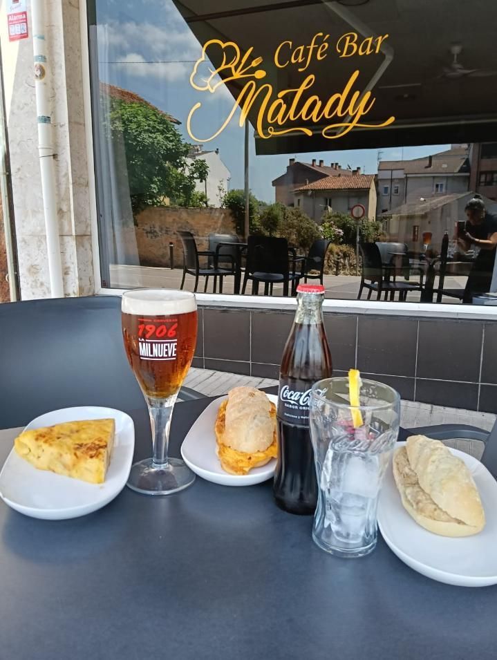 Mesa con comida y bebidas fuera de una cafetería: tortilla, sándwich, cerveza, Coca-Cola y vaso de agua. 