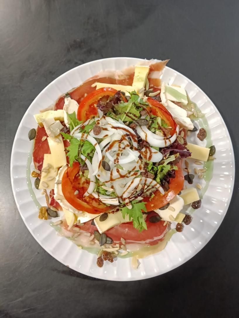 Plato de comida: prosciutto, queso, rodajas de tomate, cebolla, verduras, semillas, glaseado balsámico.