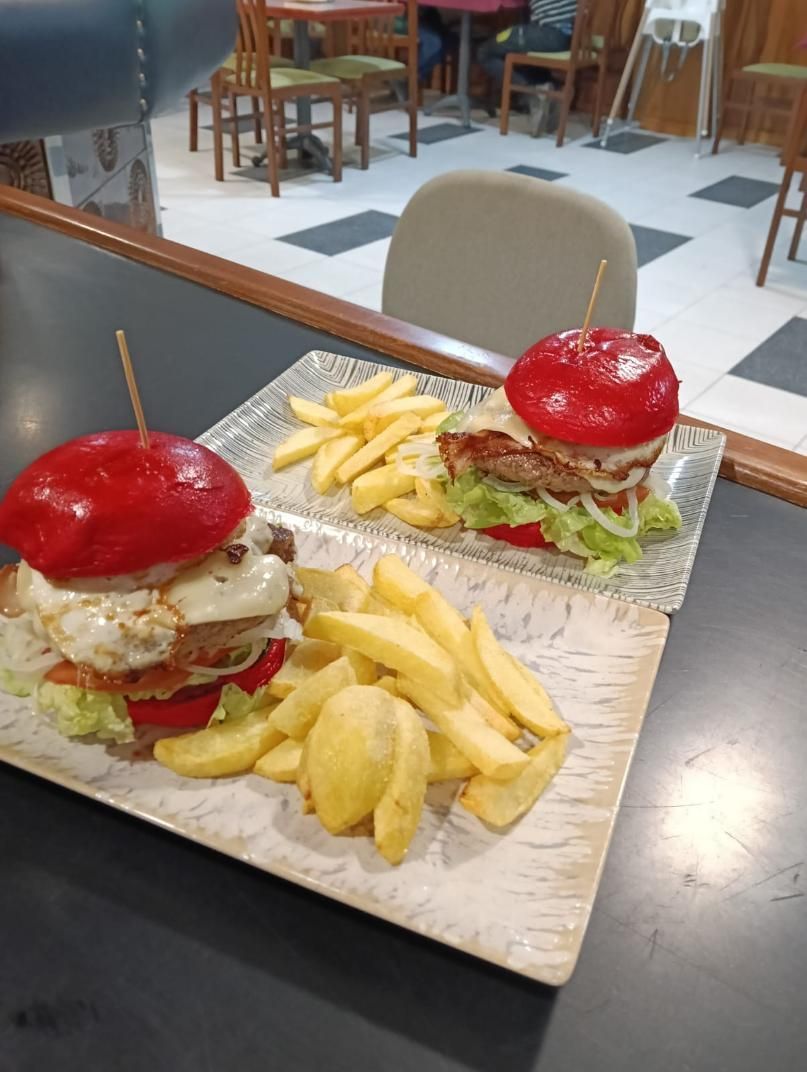 Dos hamburguesas de pan rojo con papas fritas en platos cuadrados, sobre una mesa en un restaurante.