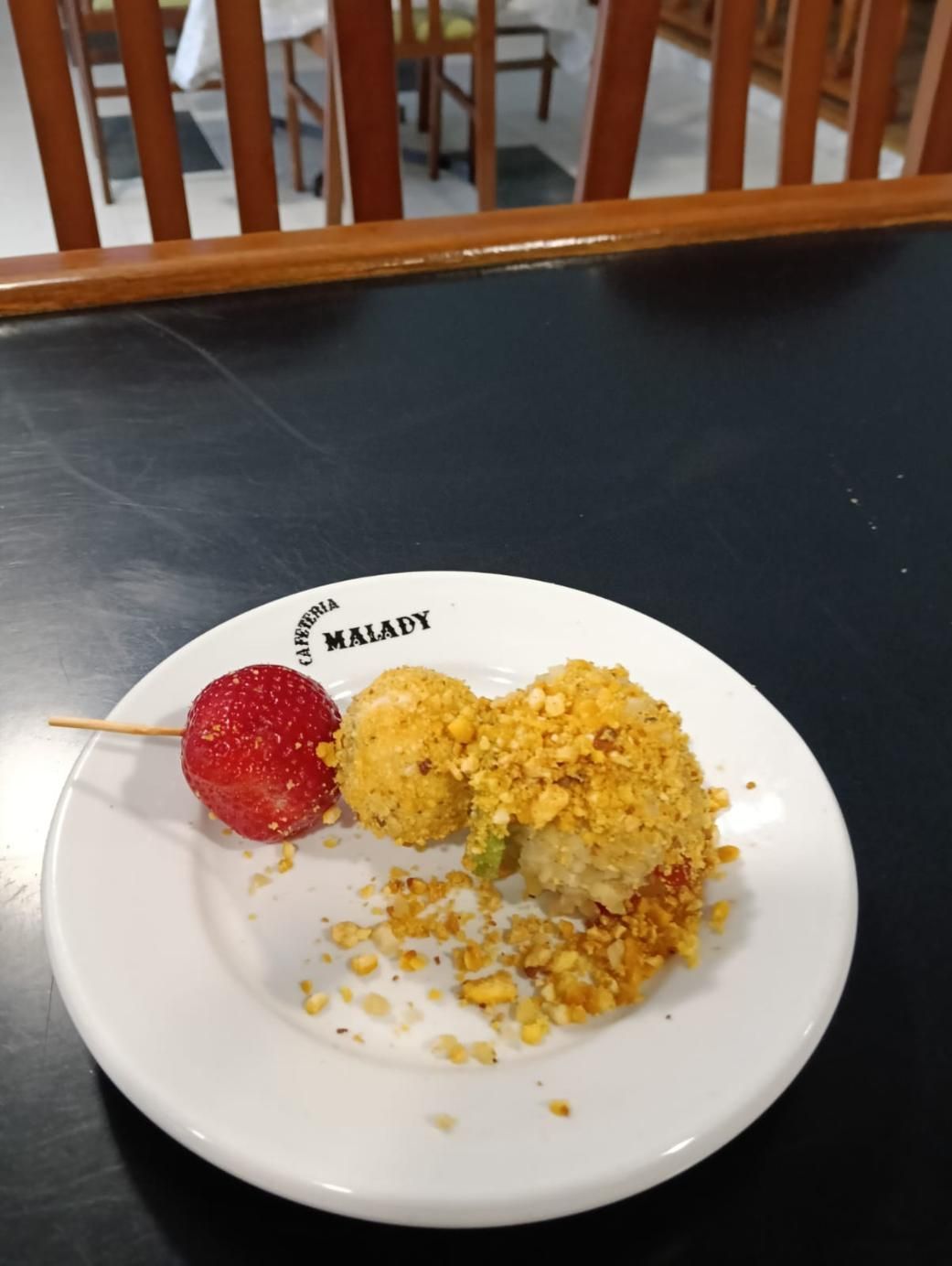 Un plato con una brocheta de fresa, dos bolas fritas y migas sobre una mesa negra.