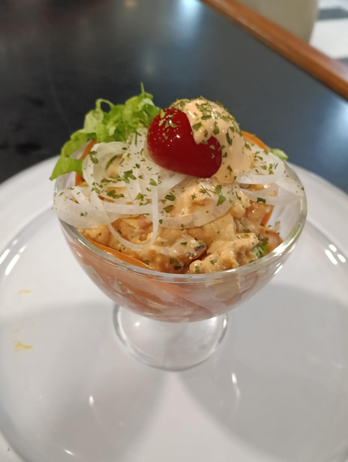 Cóctel de camarones en copa con salsa, cebolla y cereza, servido en un plato blanco.