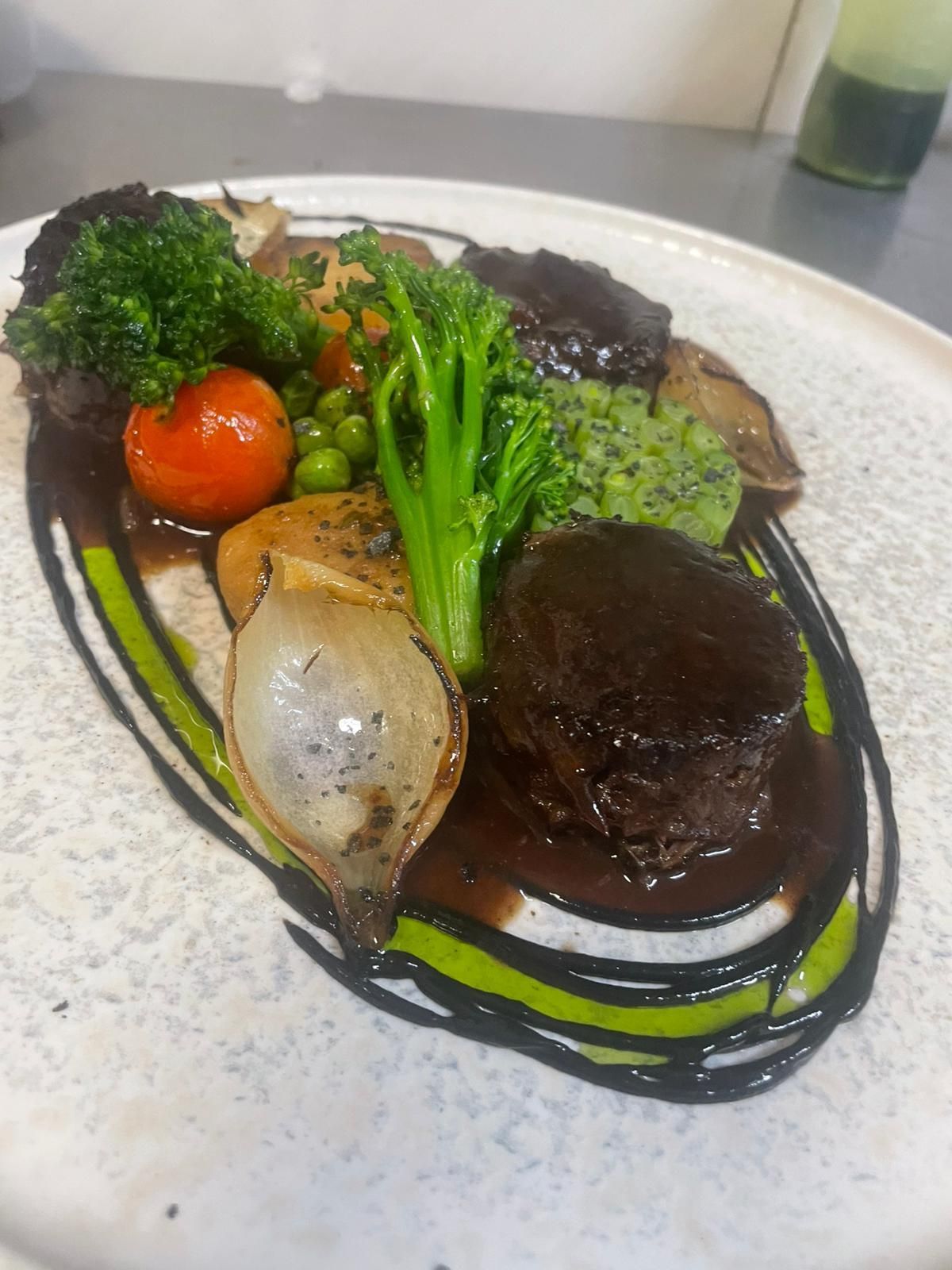 Un plato gourmet que incluye medallones sellados con salsa marrón, brócoli, cebolla asada, tomate cherry y puré de hierbas.