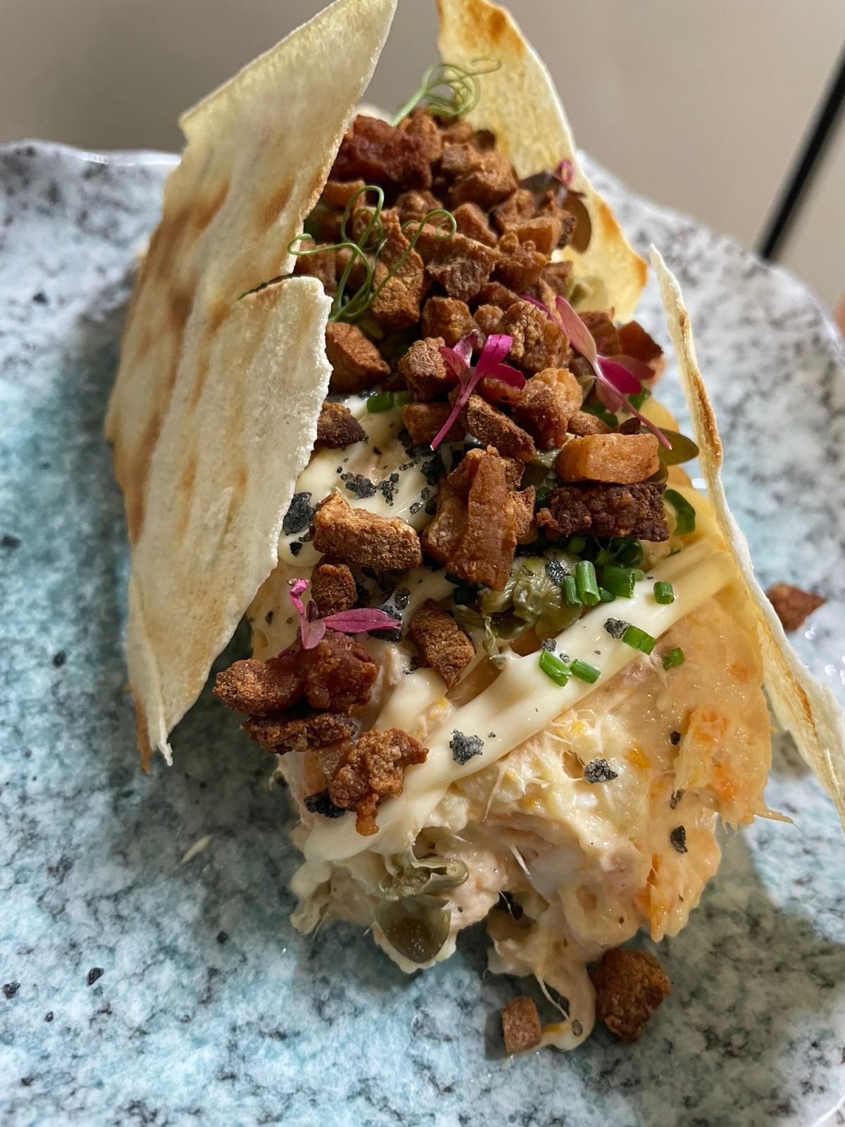 Un taco con una tortilla crujiente, relleno cremoso, cubierto con trocitos crujientes