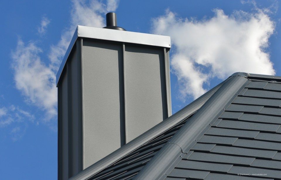 Grauer Schornstein an einem Haus mit grauem Dach vor blauem Himmel mit weißen Wolken.