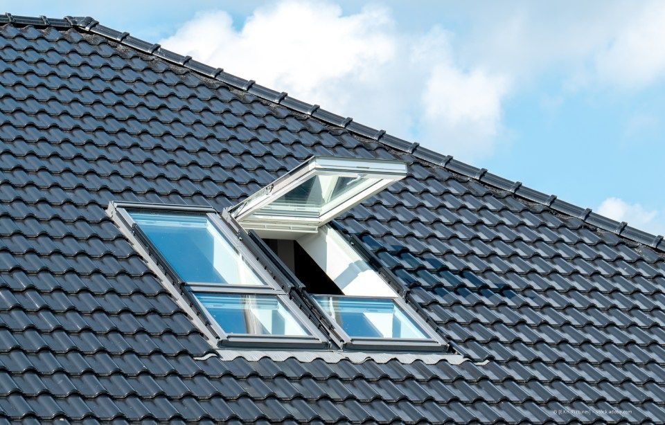 Oberlichter auf einem dunklen Ziegeldach, eines davon geöffnet, vor blauem Himmel.