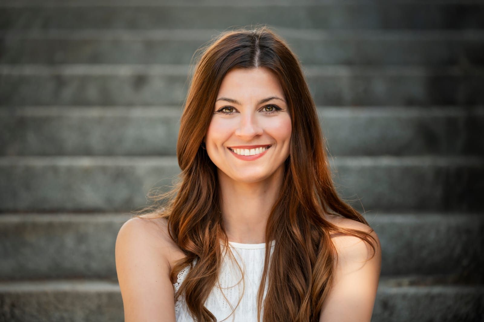 Eine Frau mit langen braunen Haaren lächelt, sitzt auf einer Steintreppe im Freien und trägt ein weißes Oberteil.