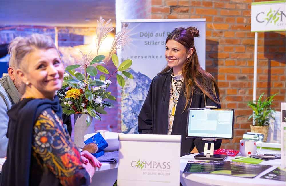 Bei einer Tagung stehen zwei Frauen nebeneinander an einem Tisch.
