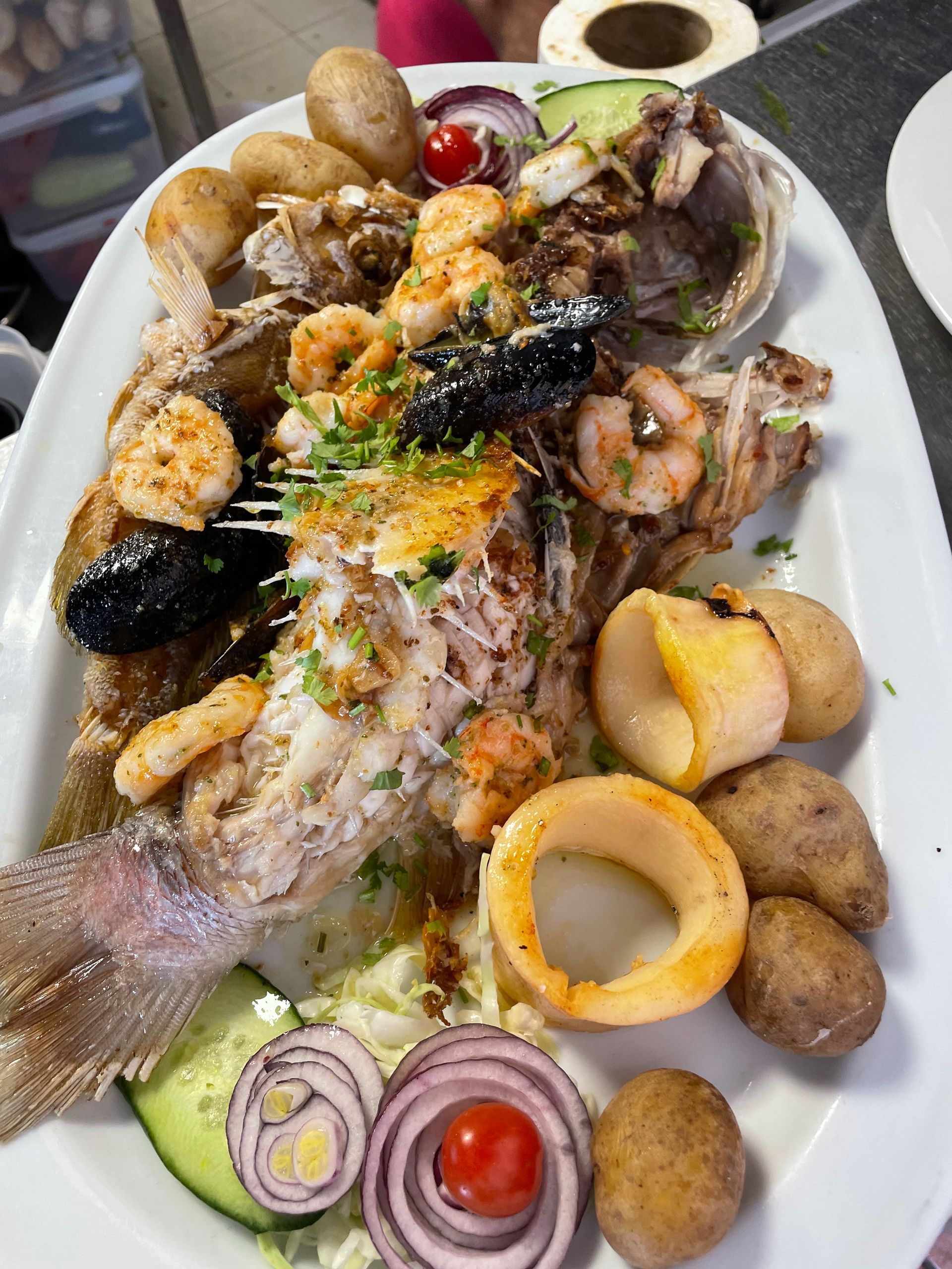 Un plato blanco cubierto con pescado, camarones, papas, cebollas y otras verduras.